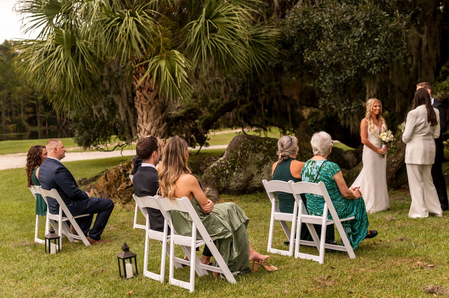 Small outdoor wedding ceremony with guests seated on white chairs under palm trees in a garden setting.