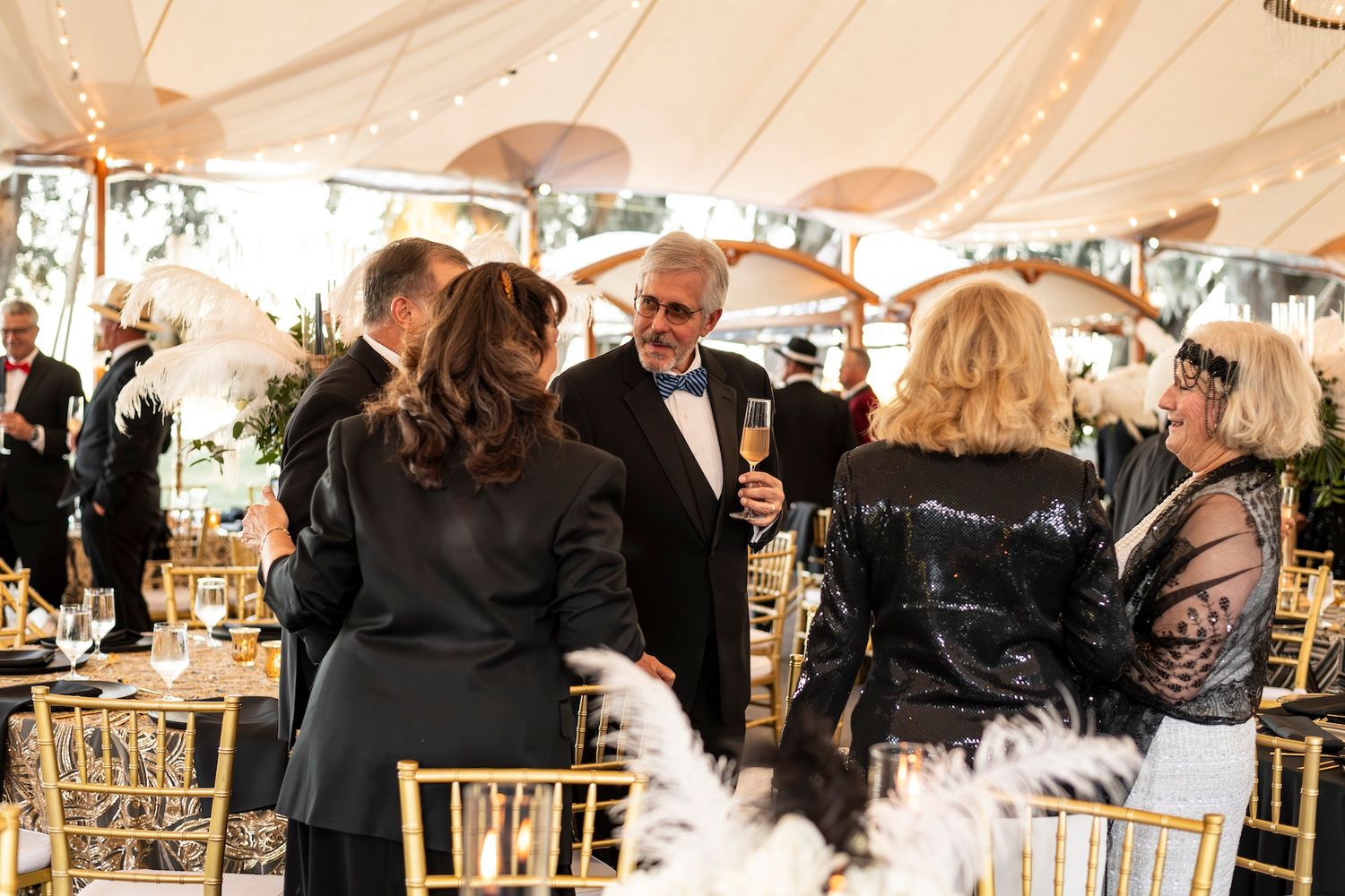 Guests mingle and converse at an elegant formal event with gold chiavari chairs under a tented reception space.