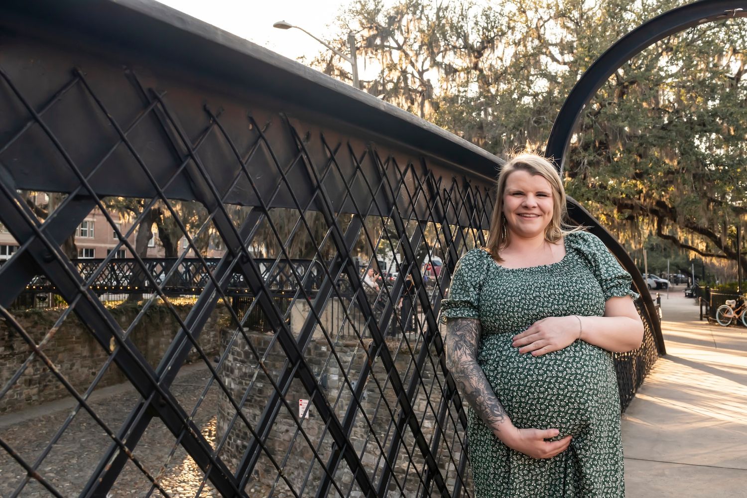 Expectant woman in green patterned dress stands near a decorative metal lattice fence in warm evening sunlight.