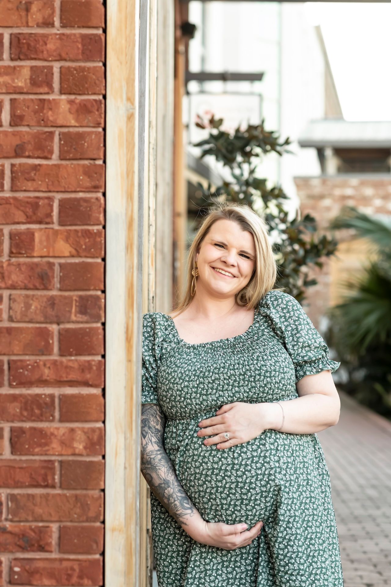 Pregnant woman wearing a green floral dress poses against brick wall for maternity photos at sunset.