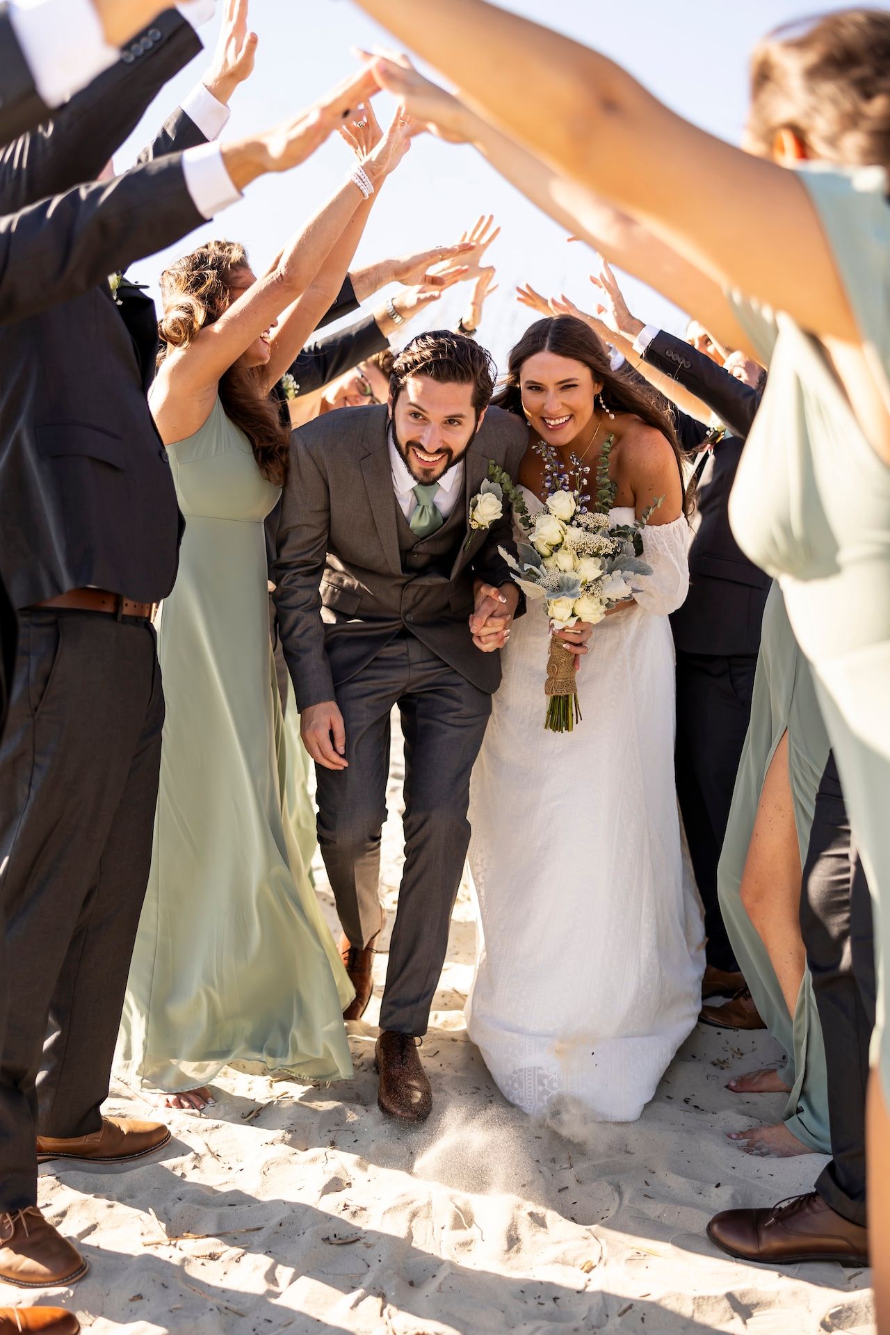 A couple walks through a tunnel of wedding guests holding their hands up to form an arch on a sunny beach.