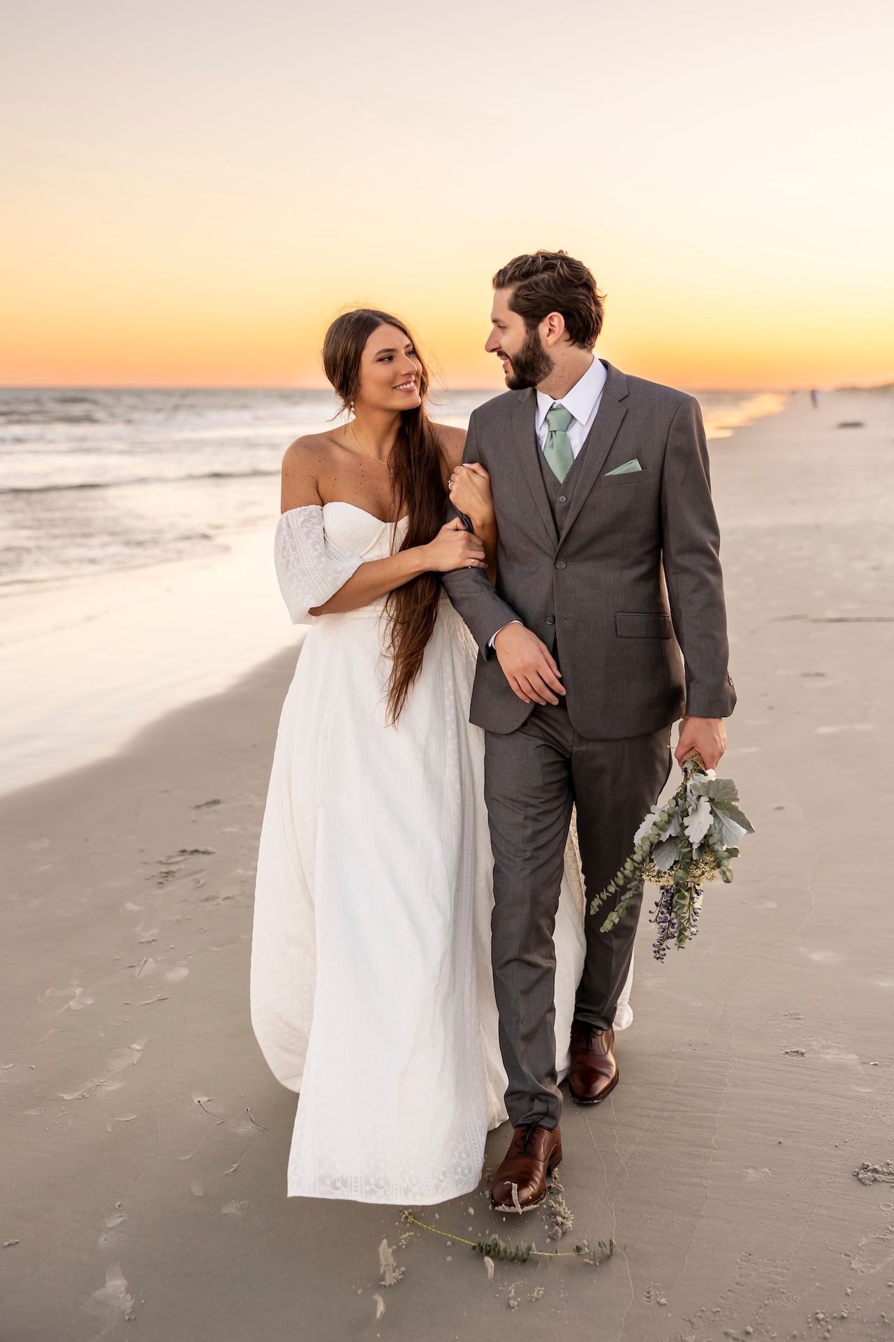 A couple in wedding attire walks hand in hand along a beach at sunset, with the groom holding a floral bouquet.