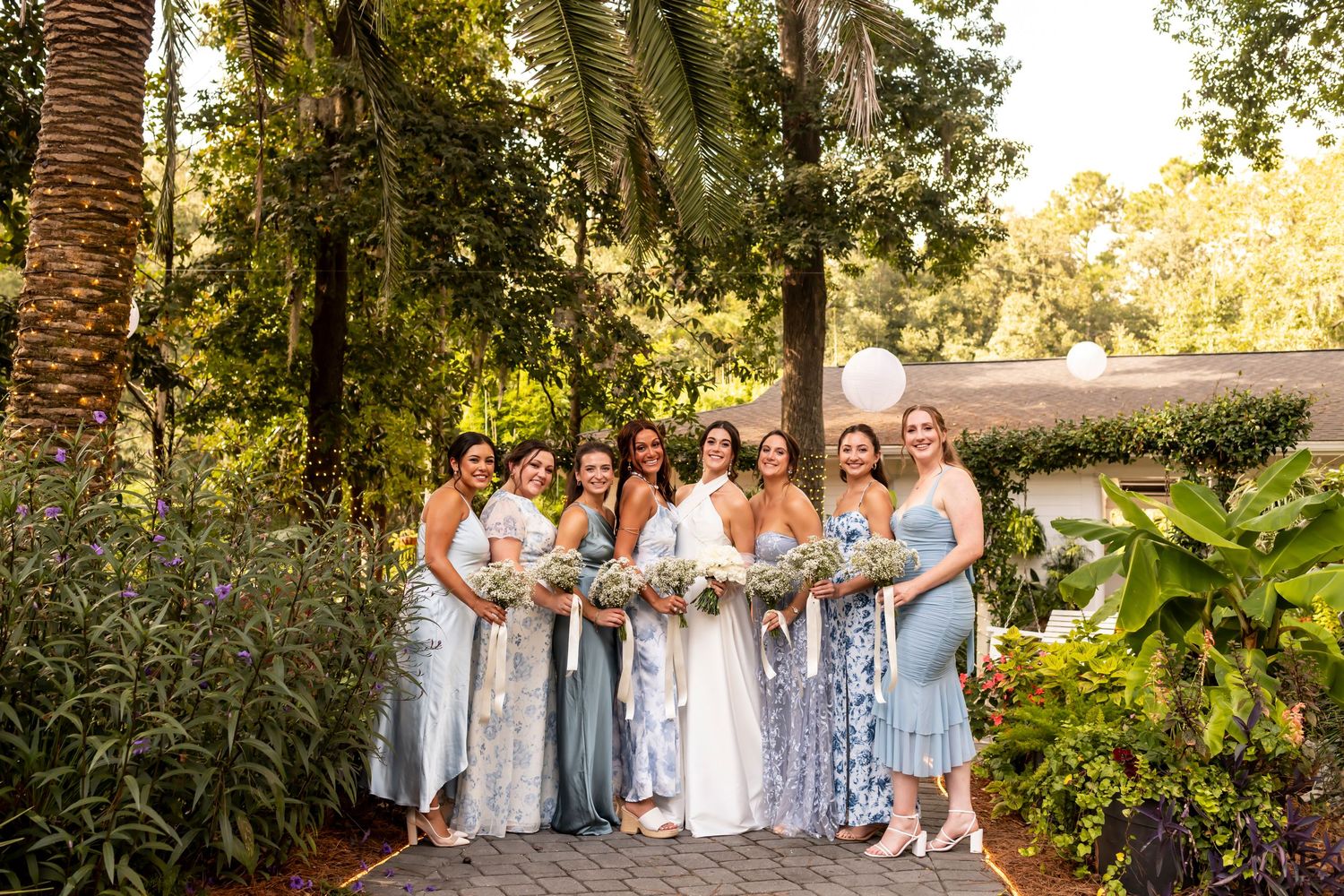 Wedding party in blue and white dresses poses on garden path surrounded by lush foliage.