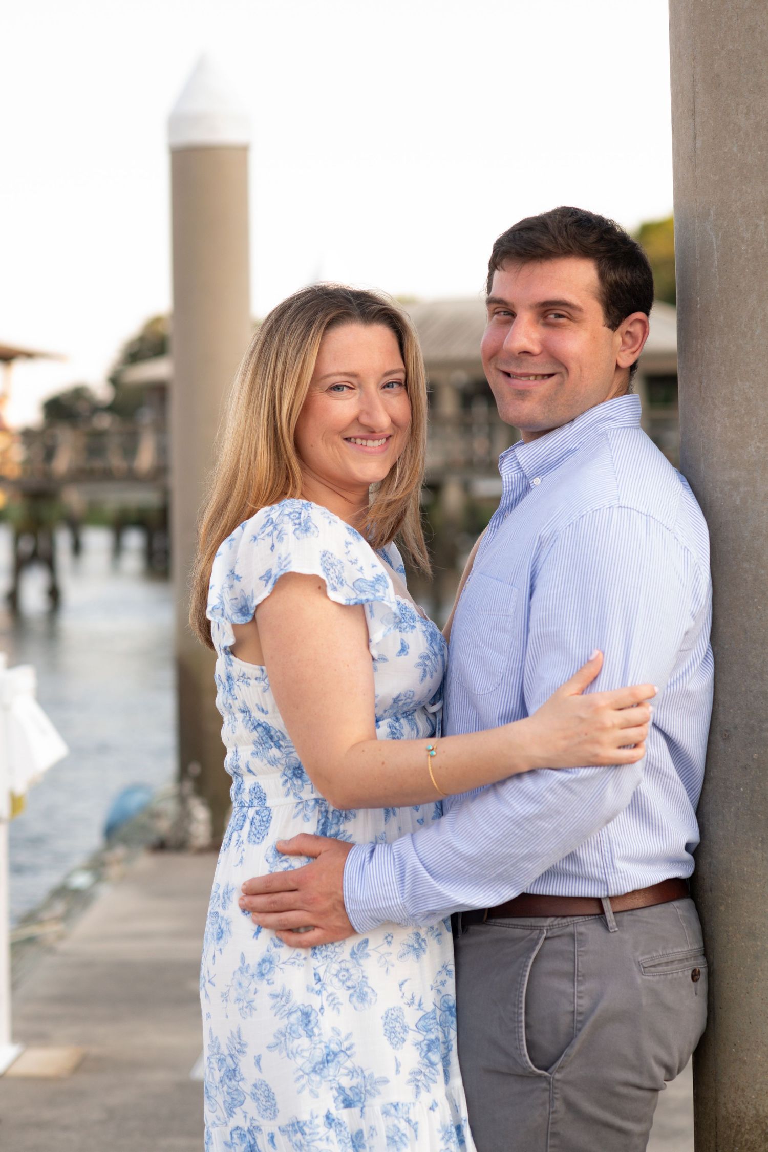 A couple in coordinated blue and white outfits share romantic moments between white pillars by the waterfront.