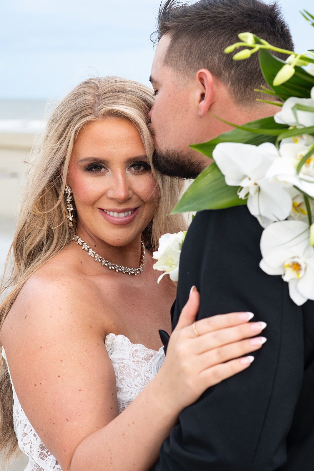A bride in a white lace dress and groom in black tuxedo embrace during their beach wedding photoshoot.