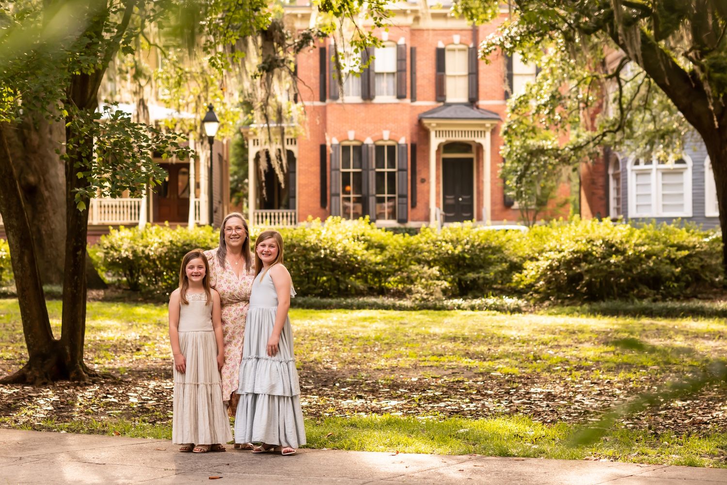 People in light-colored dresses stand together in Forsyth Park with historic brick buildings in the background.