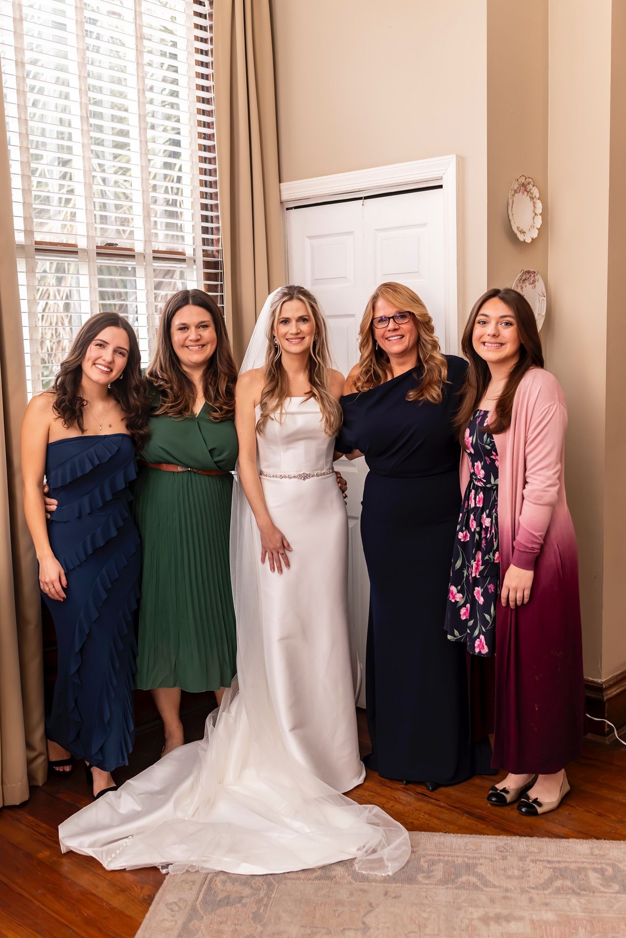 A group poses together in formal dresses at a special occasion, with a person in a flowing white gown as the centerpiece.