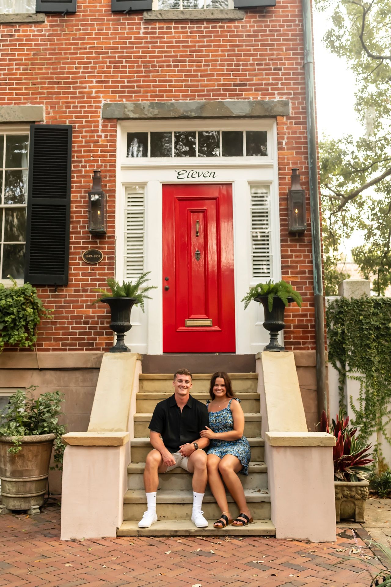 A couple sits on white stone steps in front of a historic brick townhouse with a bright red door in Savannah.