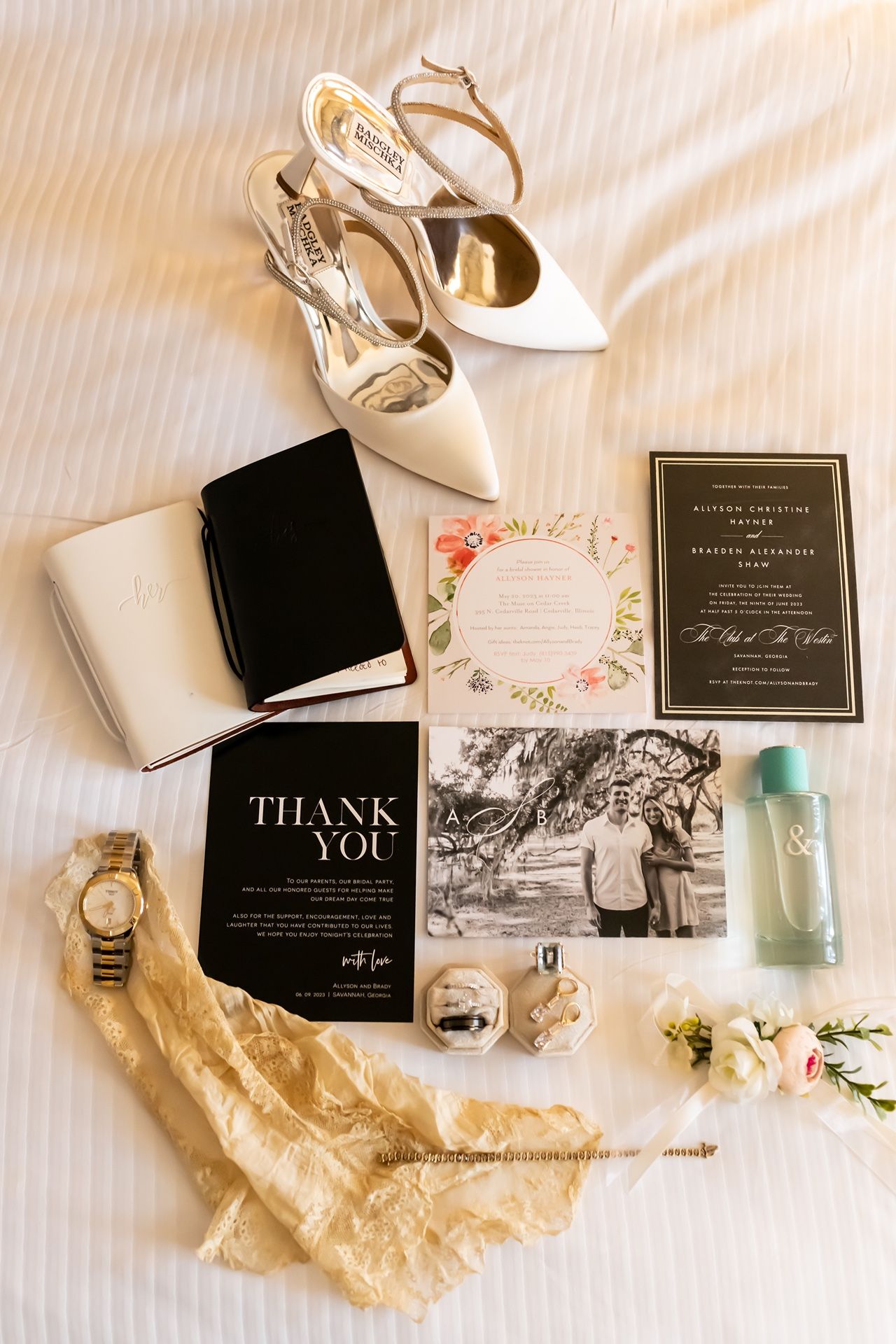 Wedding details flatlay with white heels, invitations, thank you cards, perfume bottle and accessories on white bedding.