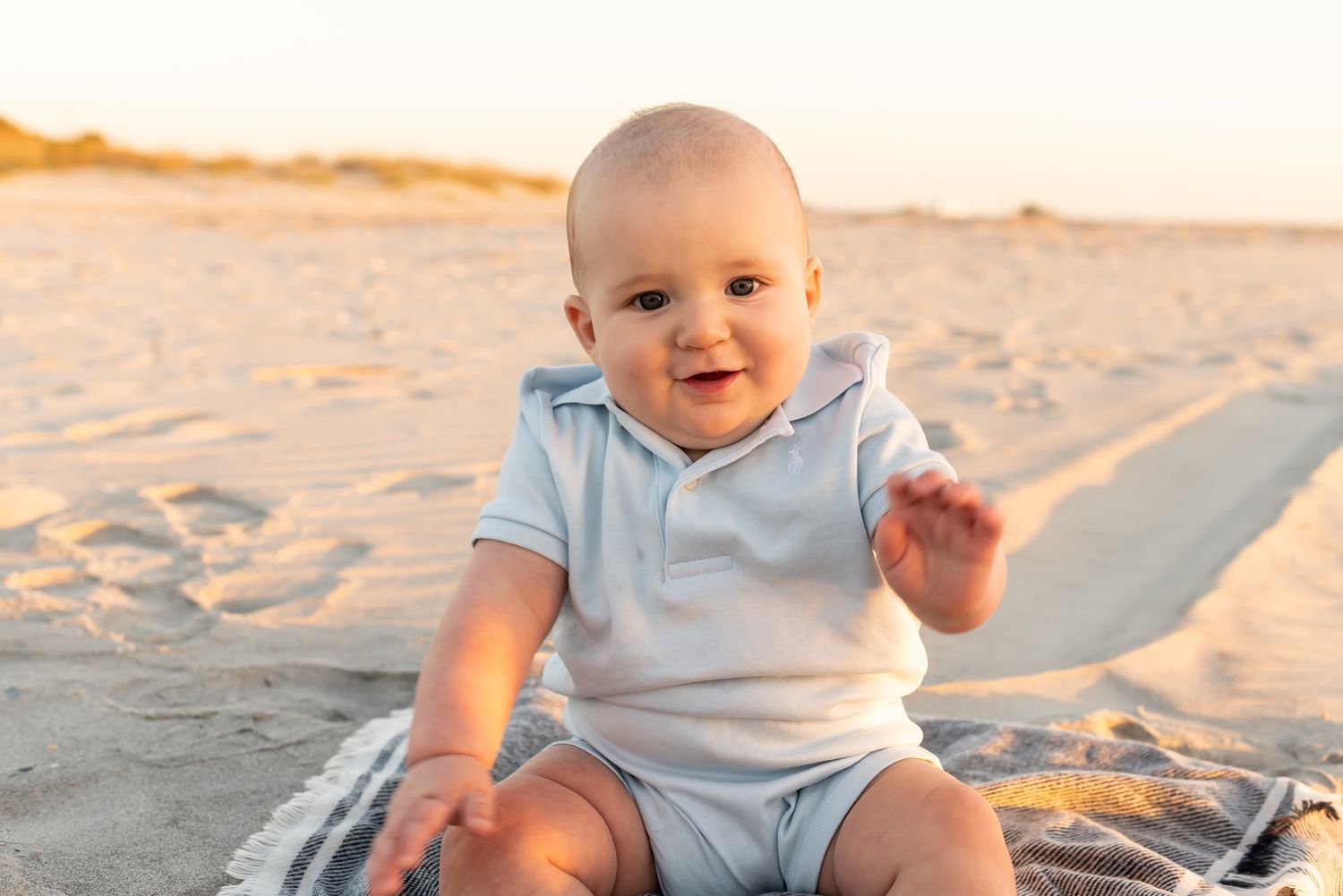 Happy baby wearing light blue onesie sits on blanket at beach during golden sunset hour.