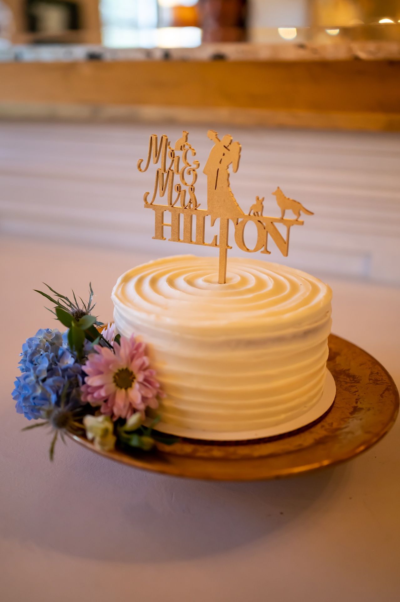 White wedding cake with swirled frosting, gold 'Mr & Mrs Hilton' topper, and blue and pink flowers on rustic wooden plate.