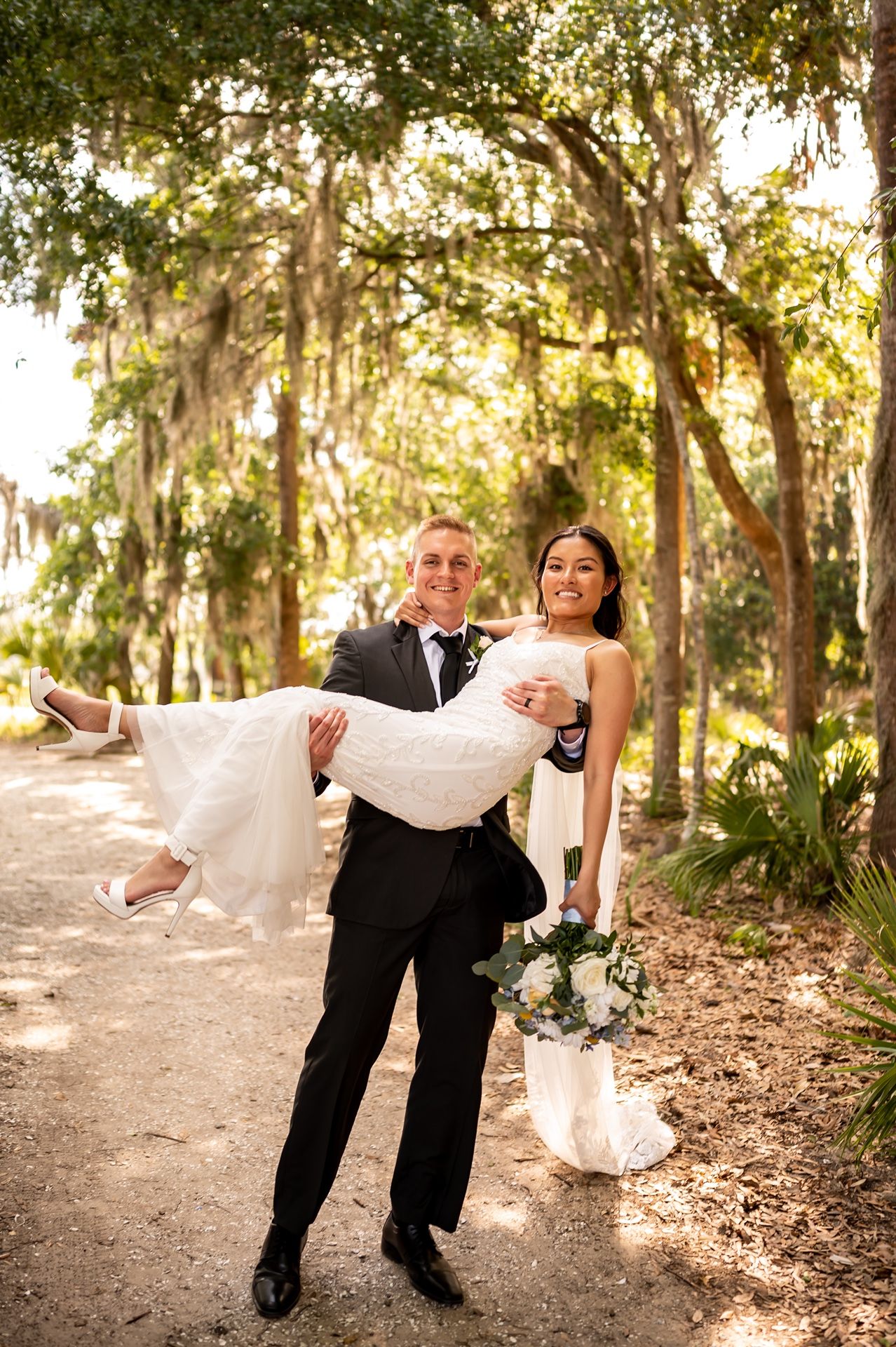 A newlywed couple share a joyful moment in Spanish moss-draped woods as the groom carries his bride.