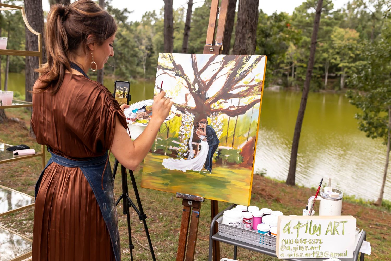 Artist painting on an easel outdoors by a lake surrounded by trees and nature.