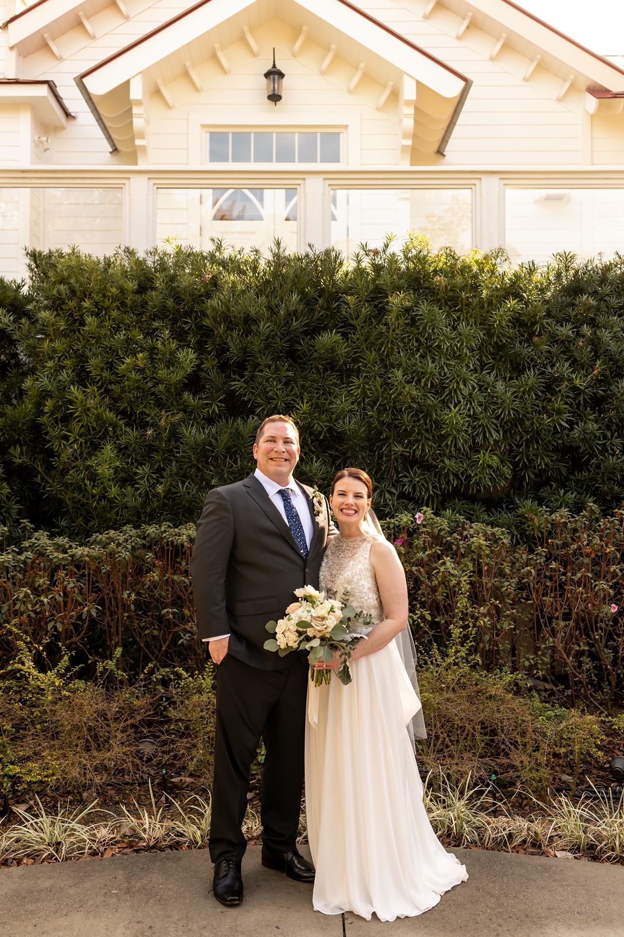 Couple poses for wedding photo in front of beige craftsman style building with lush green hedge backdrop.