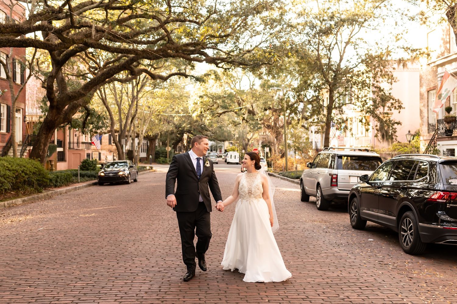 A newlywed couple walks hand in hand down a charming brick-lined street with overhanging oak trees in warm sunlight.