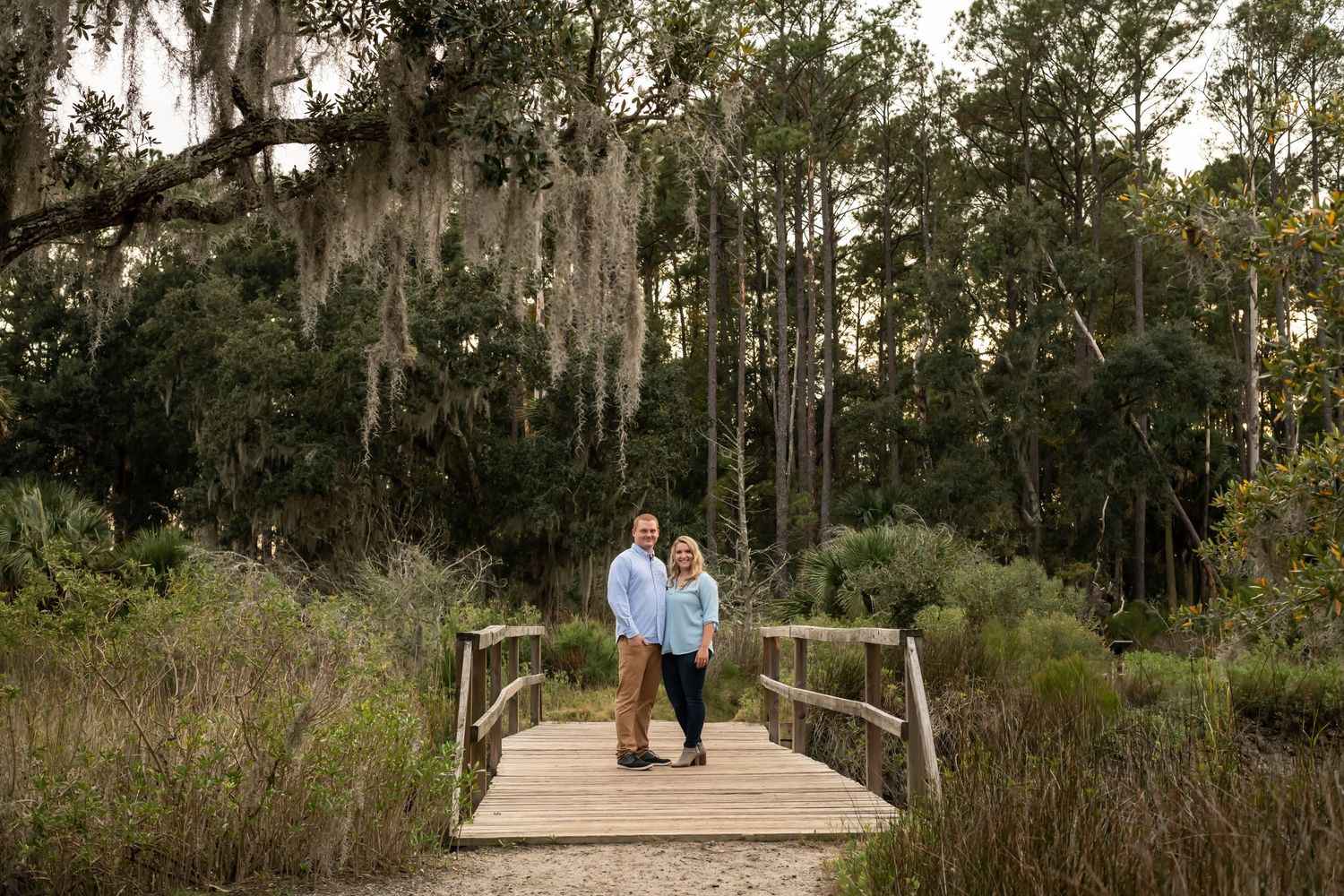 A wooden boardwalk extends through lush coastal wetlands with Spanish moss hanging from trees in the background.