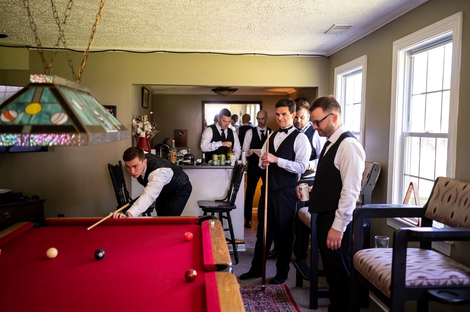 Several groomsmen in formal vests gather around a pool table in a room with a vintage-style stained glass lamp above.