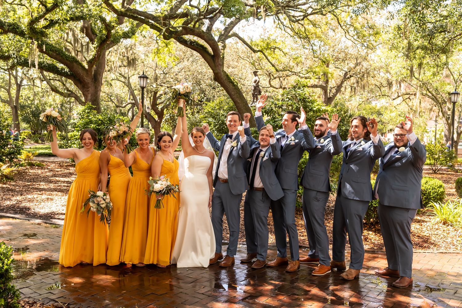 Wedding party in yellow bridesmaid dresses and gray suits poses under trees in outdoor garden setting.