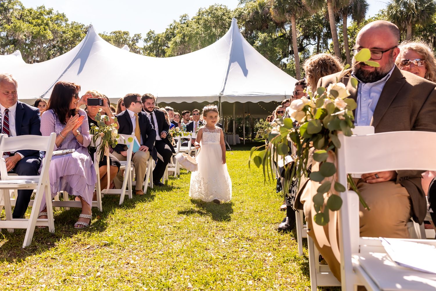 A young flower girl walks down the aisle during an outdoor wedding ceremony with white tents and sunlit grass.
