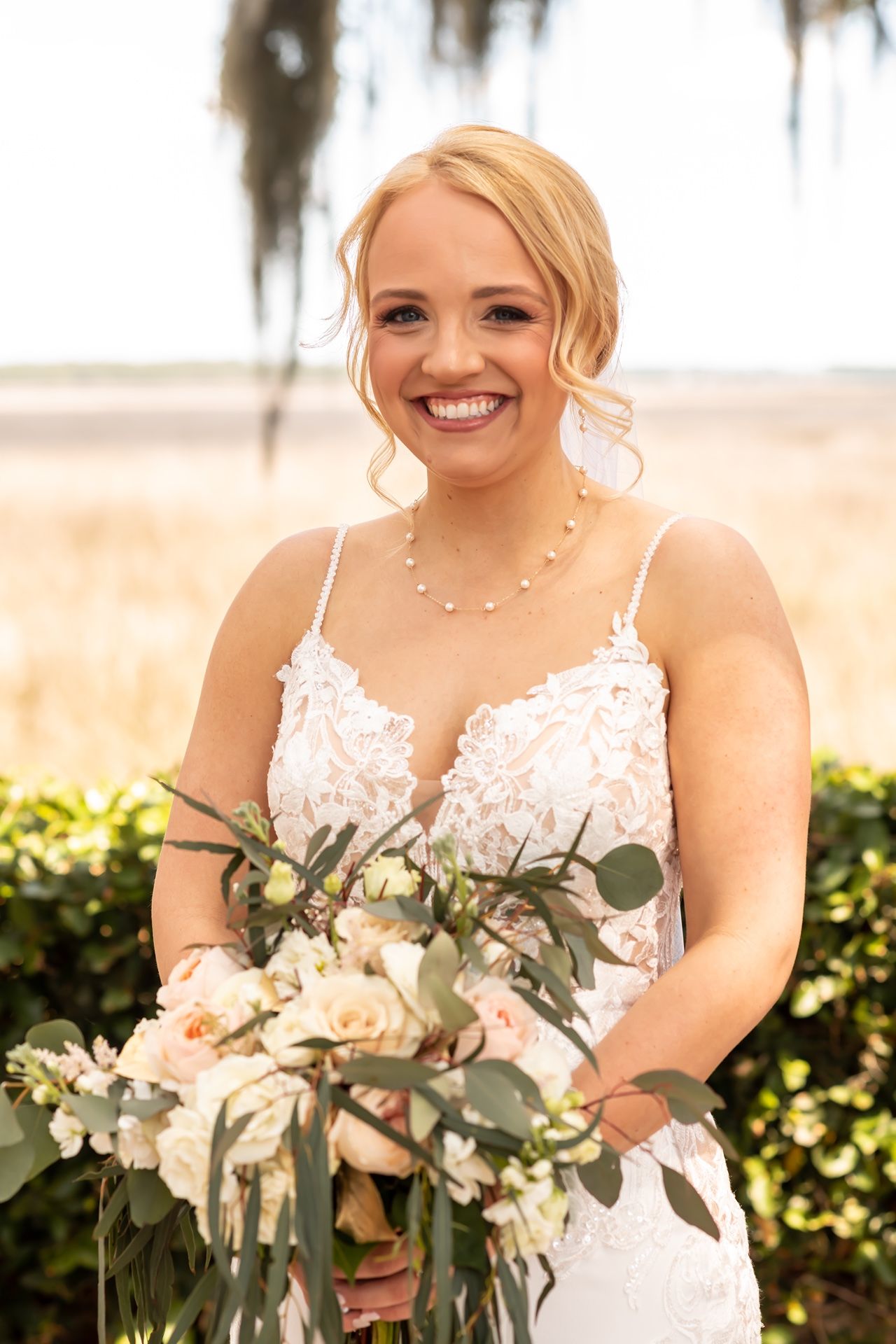 A bride wearing a white lace wedding dress smiles while holding a pink and green eucalyptus bouquet outdoors.