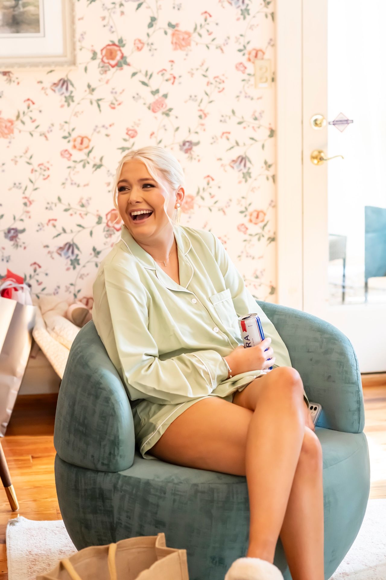 An individual wearing a cream-colored sweater sits comfortably on a teal armchair in a room with floral wallpaper.