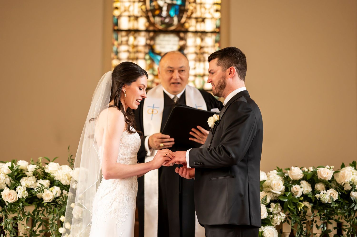 A couple exchanges rings during their wedding ceremony at an altar decorated with white floral arrangements.