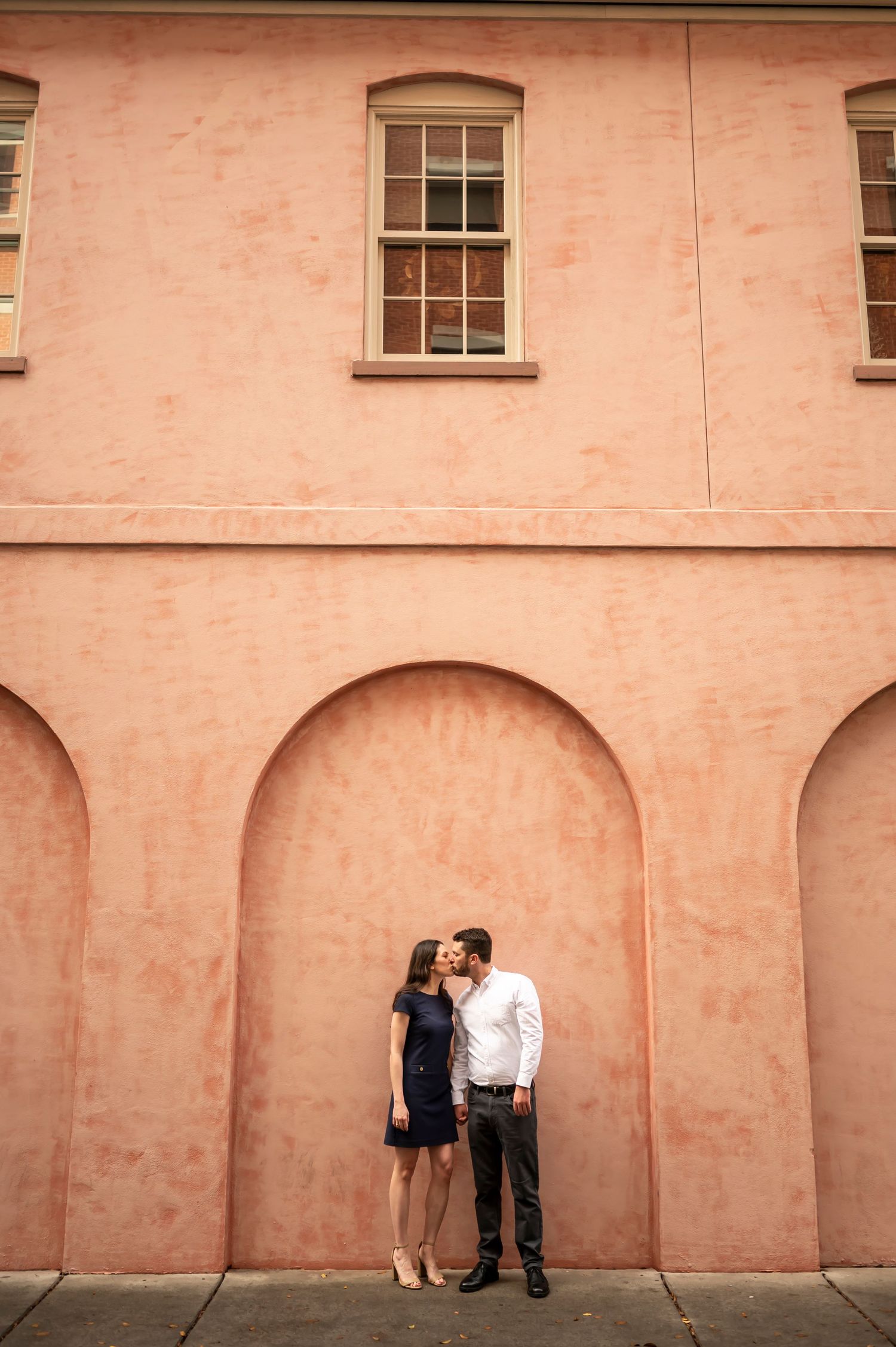Two people stand intimately against a peach-colored building with arched architectural details and windows above.