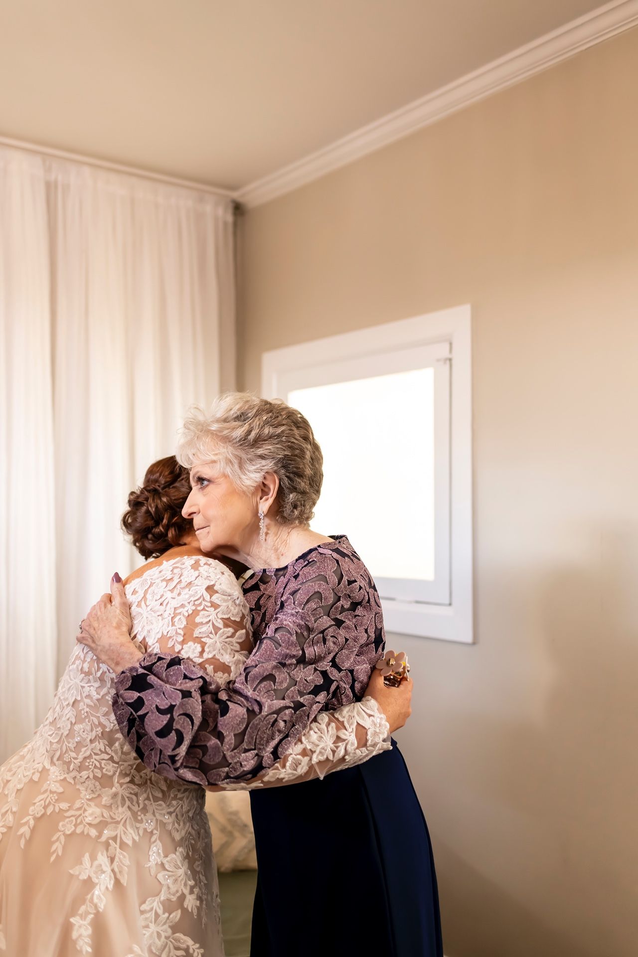 Two figures embrace in a tender moment against soft beige walls and white curtains.