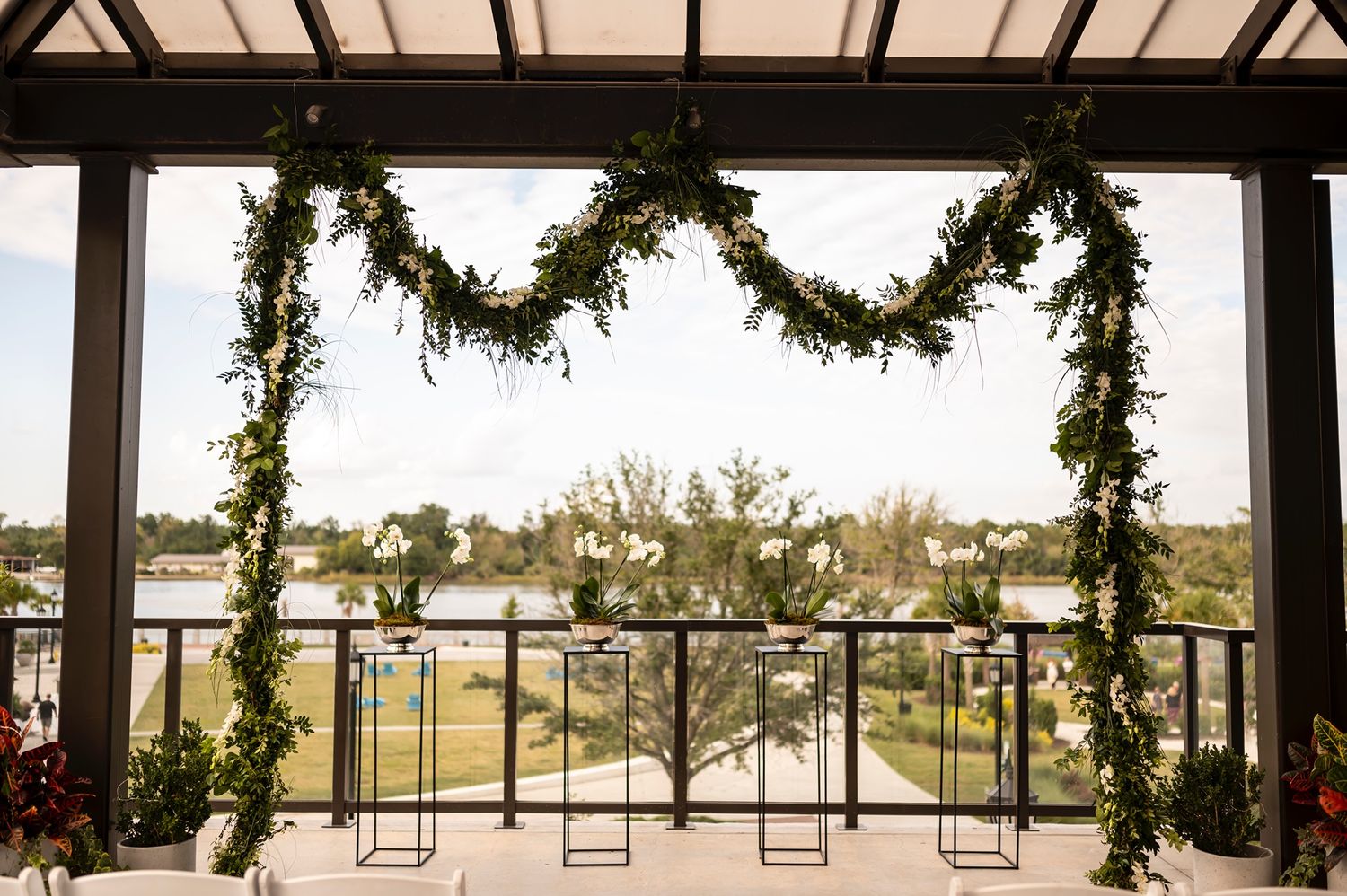 Elegant wedding arch with greenery garland overlooks waterfront venue through large glass windows.