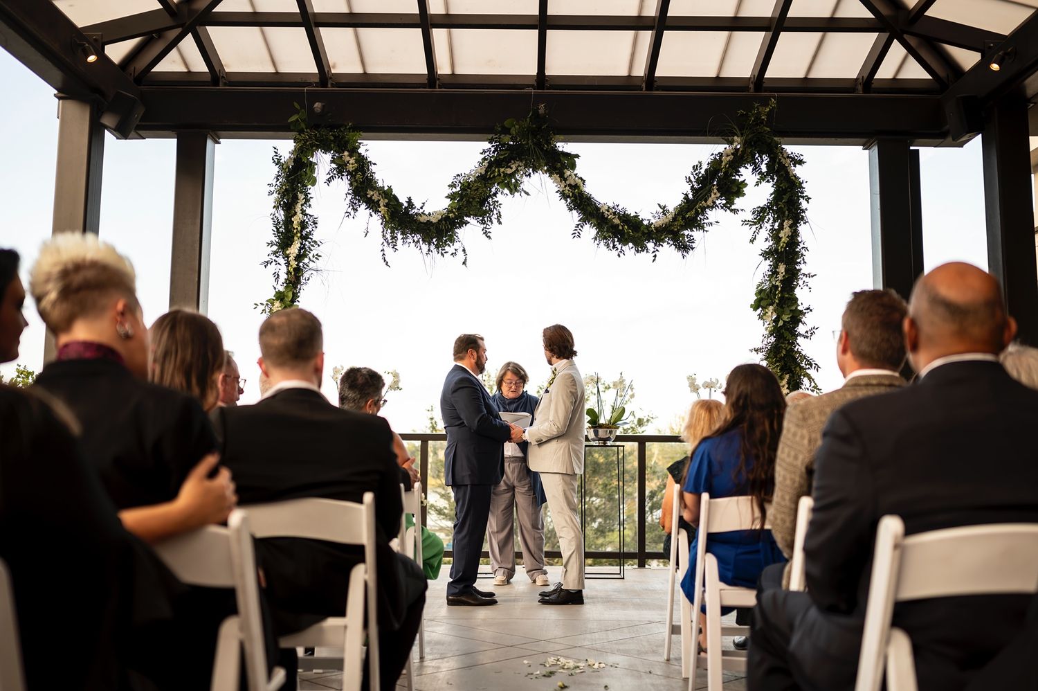 Wedding ceremony taking place under a covered pavilion decorated with greenery garlands as guests watch from white chairs.