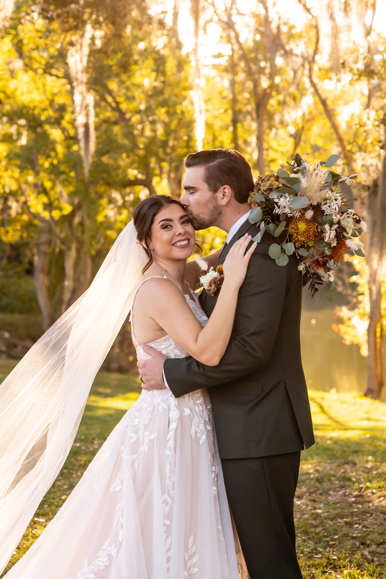 Bride and groom share romantic moment in golden sunset light with flowing veil and autumn bouquet in outdoor setting.