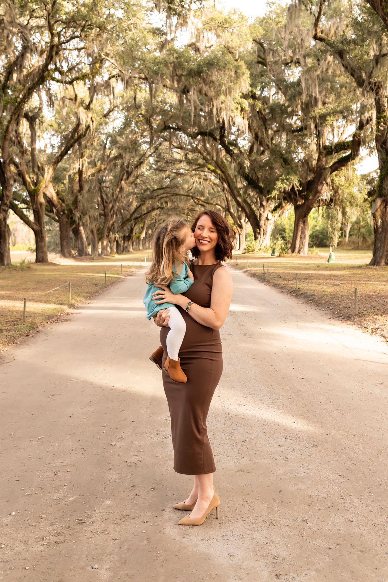 A brown-dressed figure holding a small child stands on a dirt path beneath majestic oak trees draped with Spanish moss.