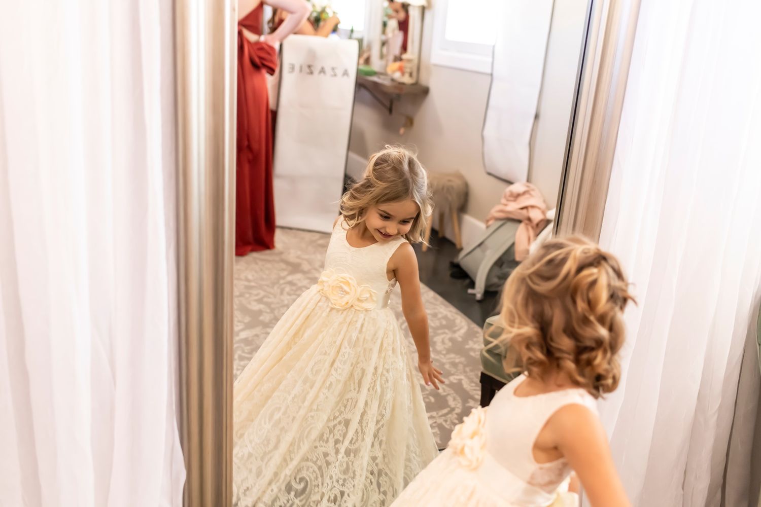 A bride in a white lacey wedding dress admires her reflection in a large mirror inside a dressing room.