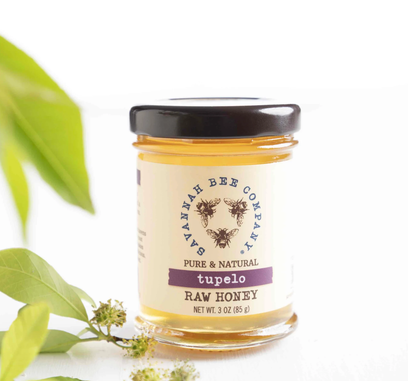 A jar of Savannah Bee Company's pure and natural tupelo raw honey displayed with green leaves and flowers in soft lighting.