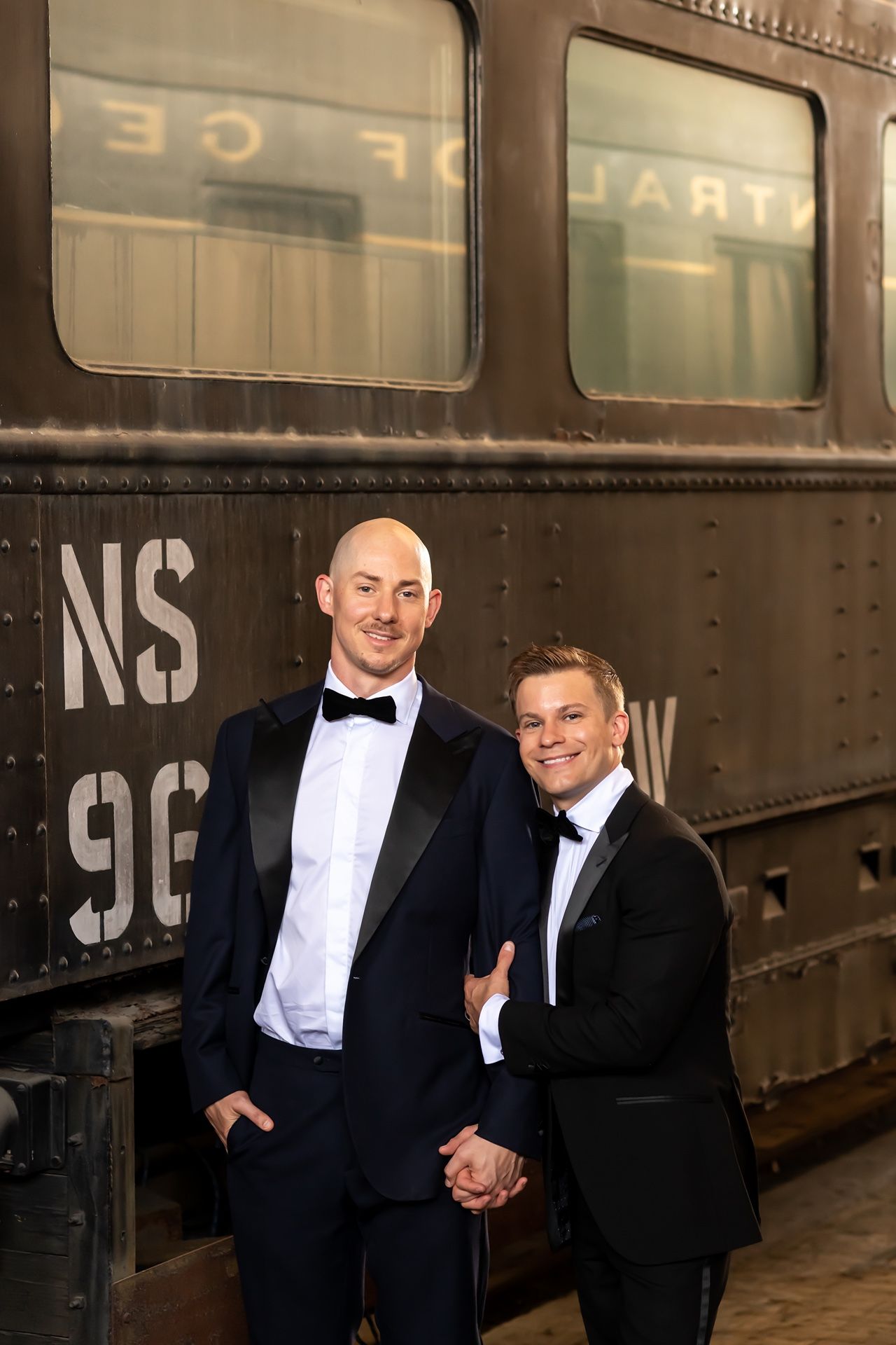Two people in formal black tuxedos pose together in front of a vintage train car.