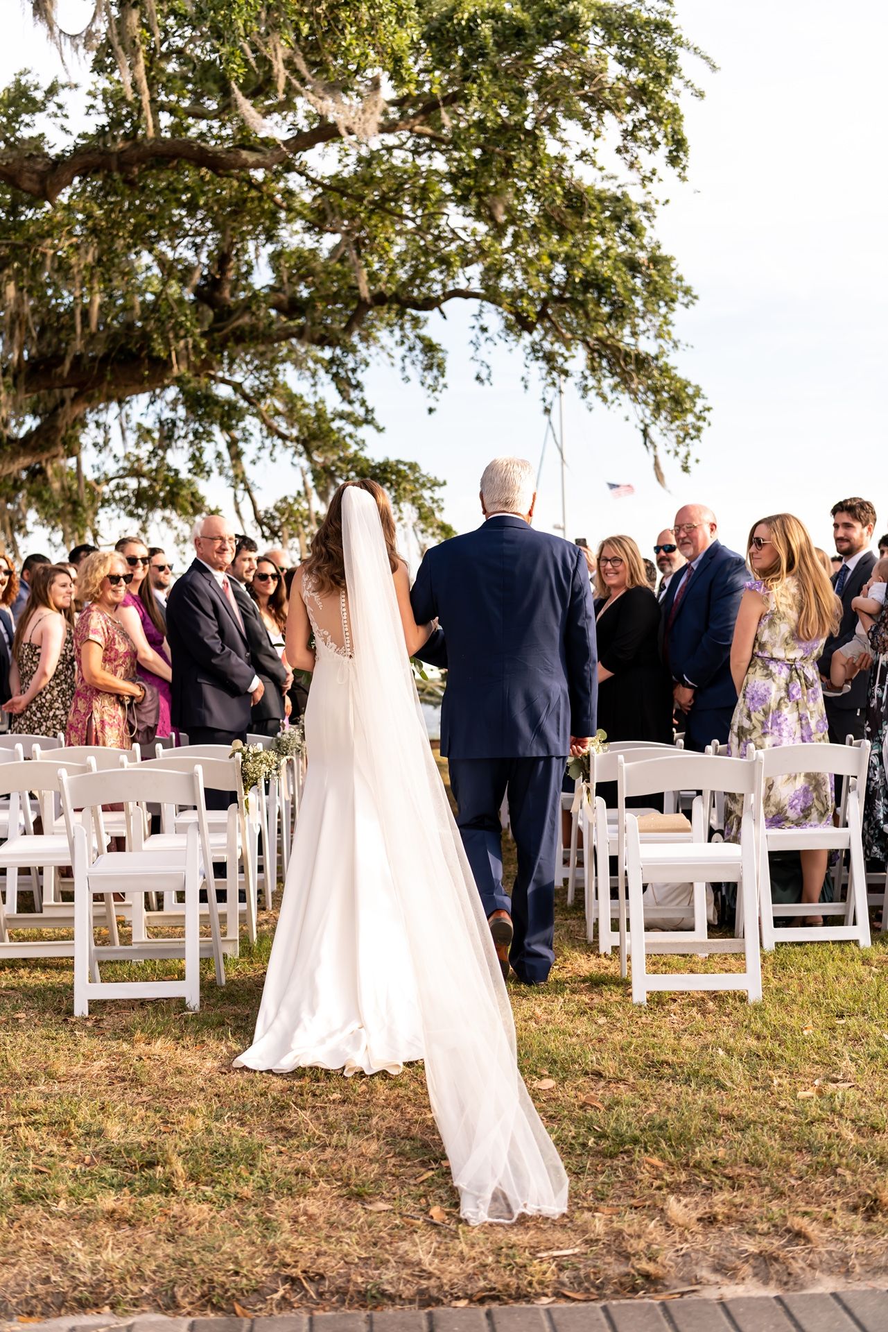 Outdoor wedding ceremony setup with white chairs under oak trees as guests gather.