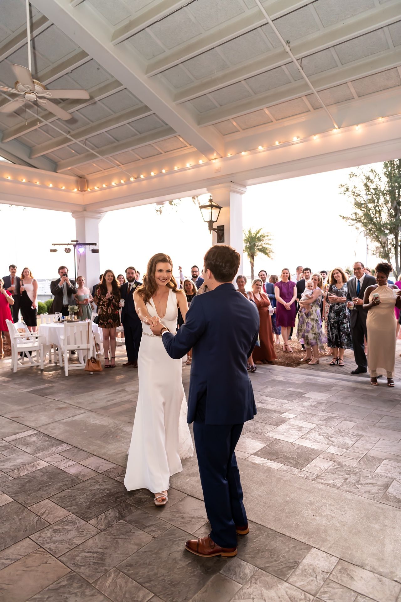 A couple performs their first dance on a stone patio under a covered pavilion surrounded by wedding guests.