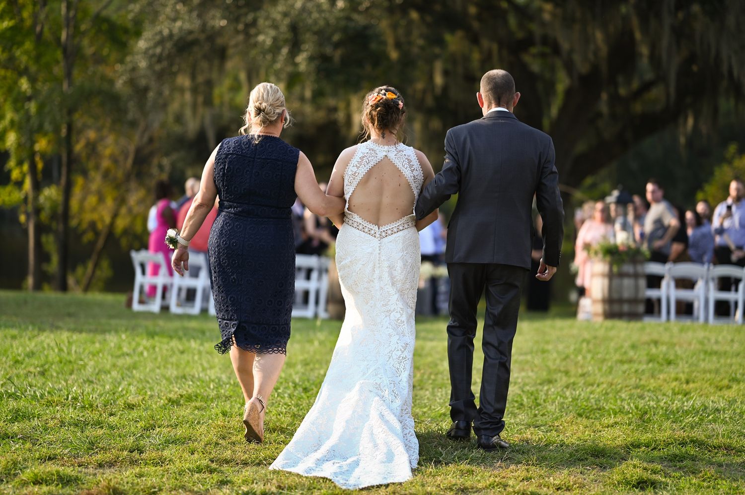 Couple walks down grassy aisle at outdoor wedding ceremony with attendant in navy dress leading the way.
