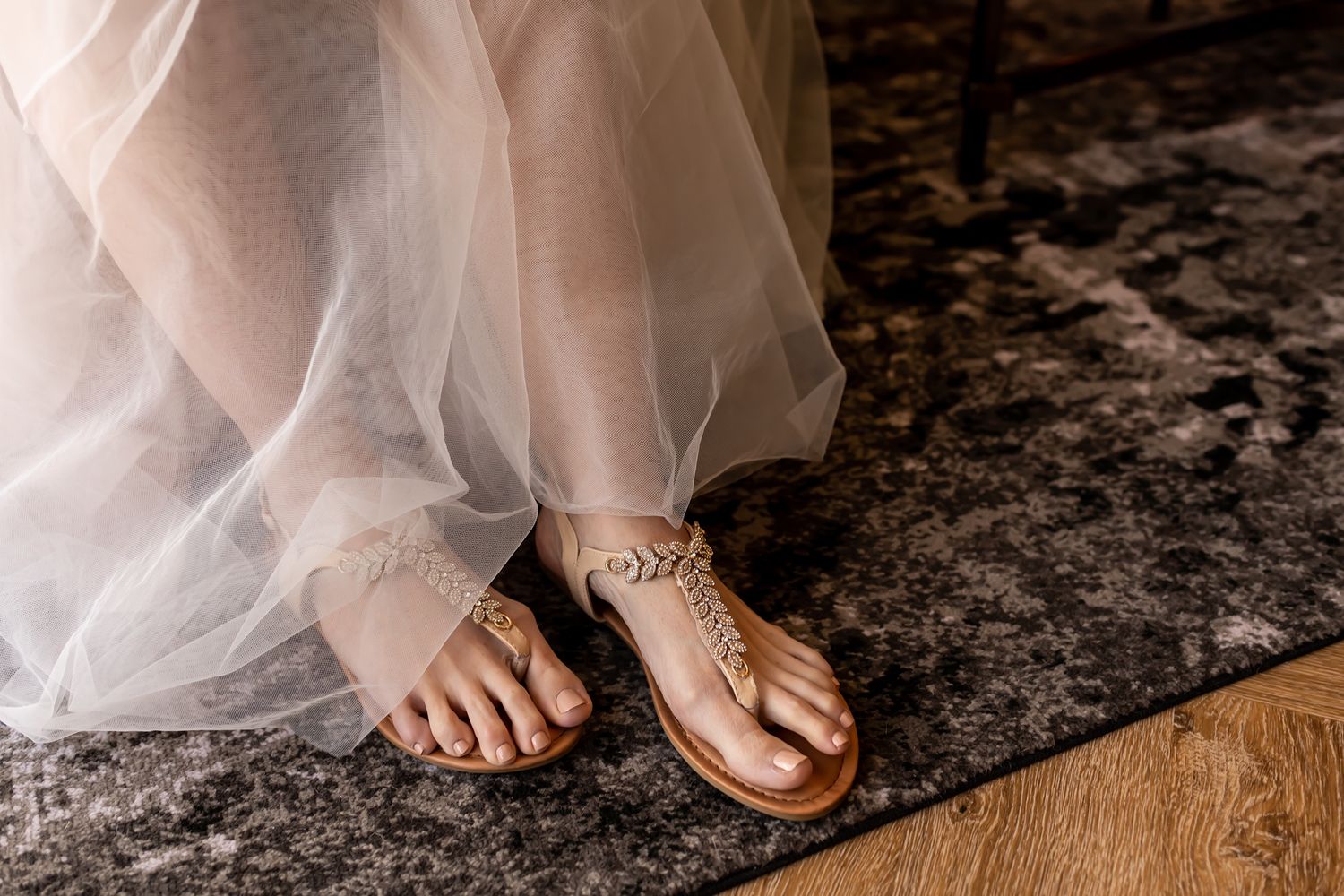 A bride wearing delicate sandals with crystal embellishments peeks out from under a flowing tulle wedding dress.