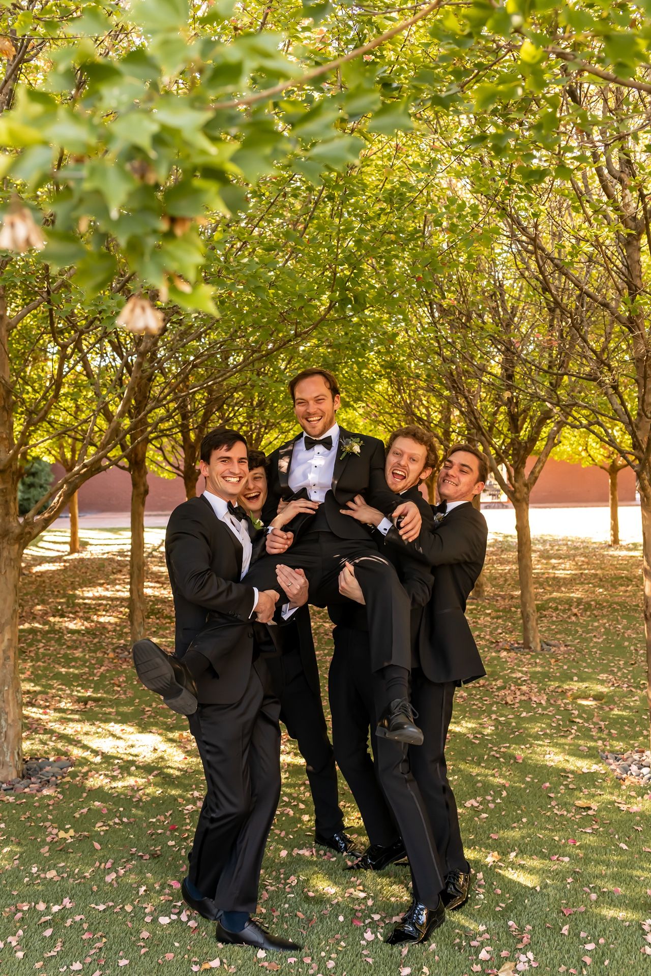 Wedding party in black tuxedos laughing together under trees with green foliage in natural lighting.