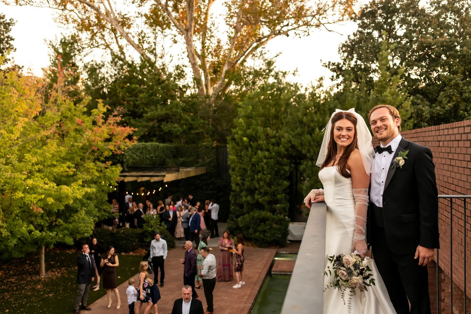 Newlyweds stand together at outdoor garden wedding venue as guests mingle on path during golden hour sunset.