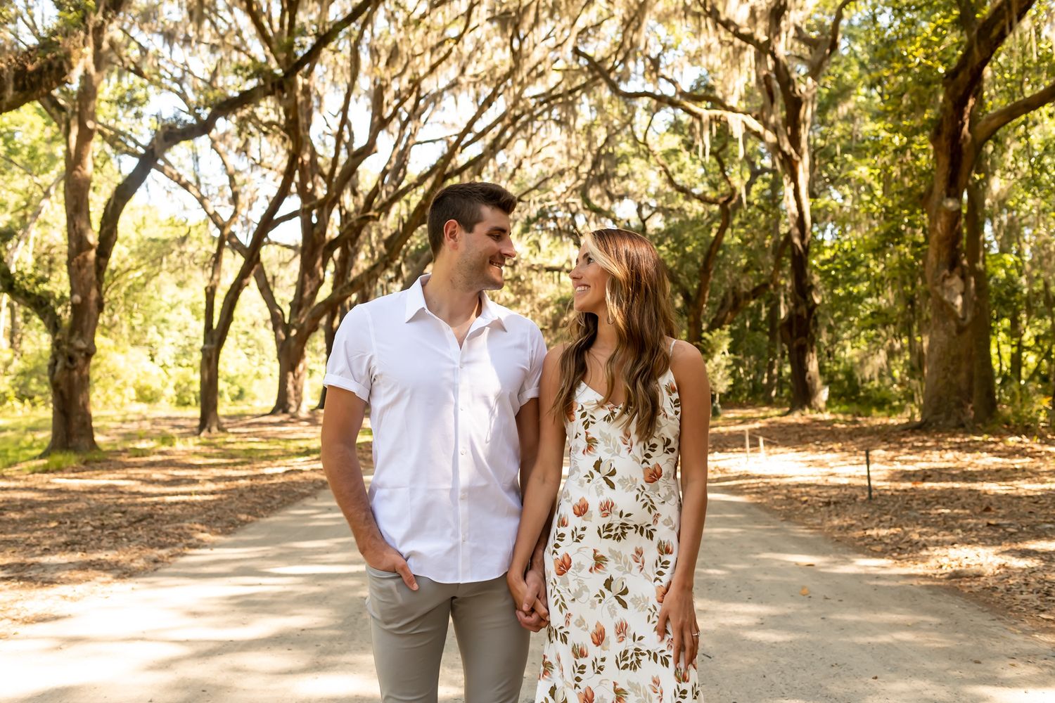 A couple walking together on a sunlit tree-lined path with spanish moss hanging from branches overhead.