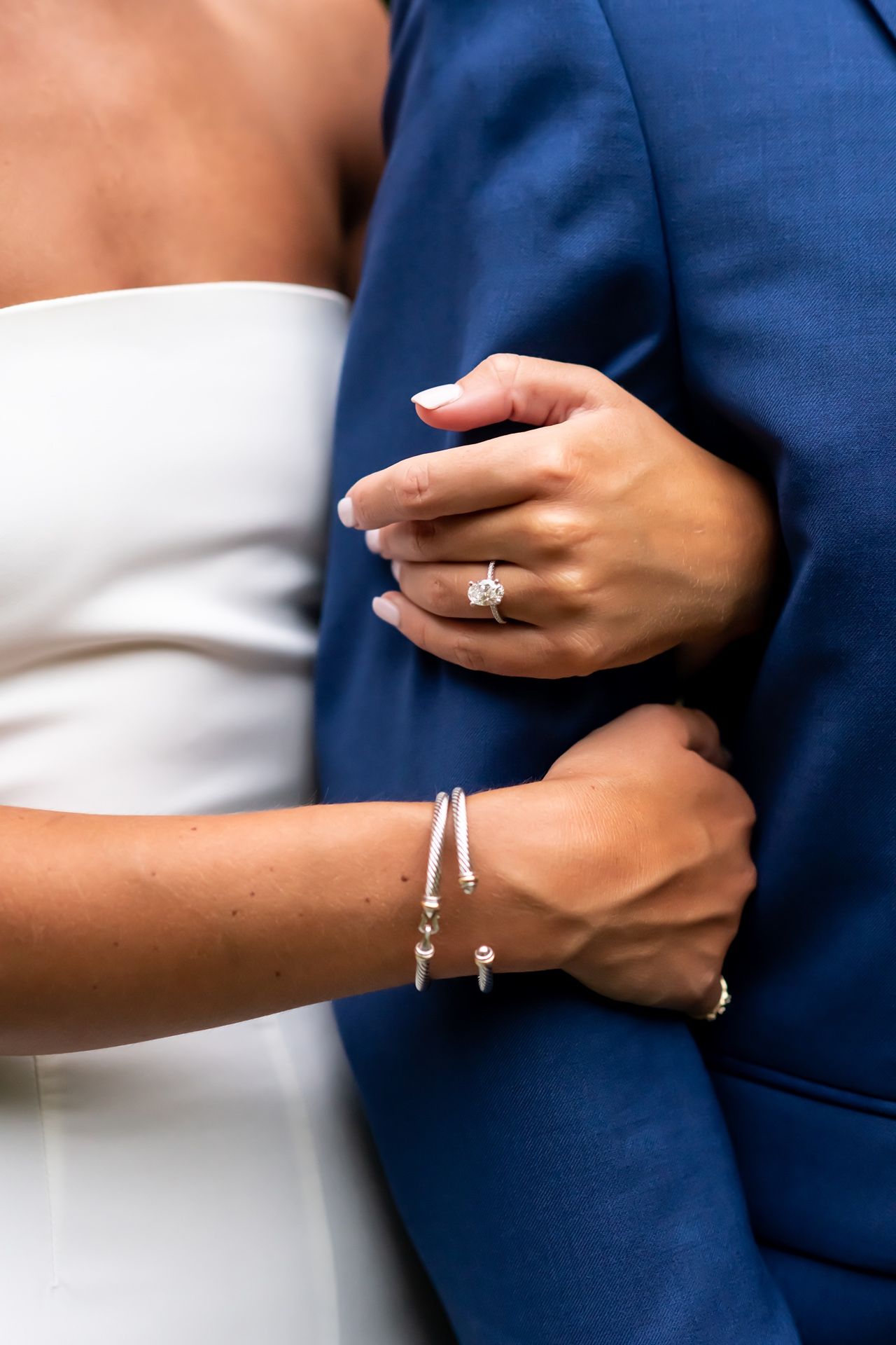 Closeup of an engagement ring and delicate bracelet jewelry worn by a couple in wedding attire embracing.