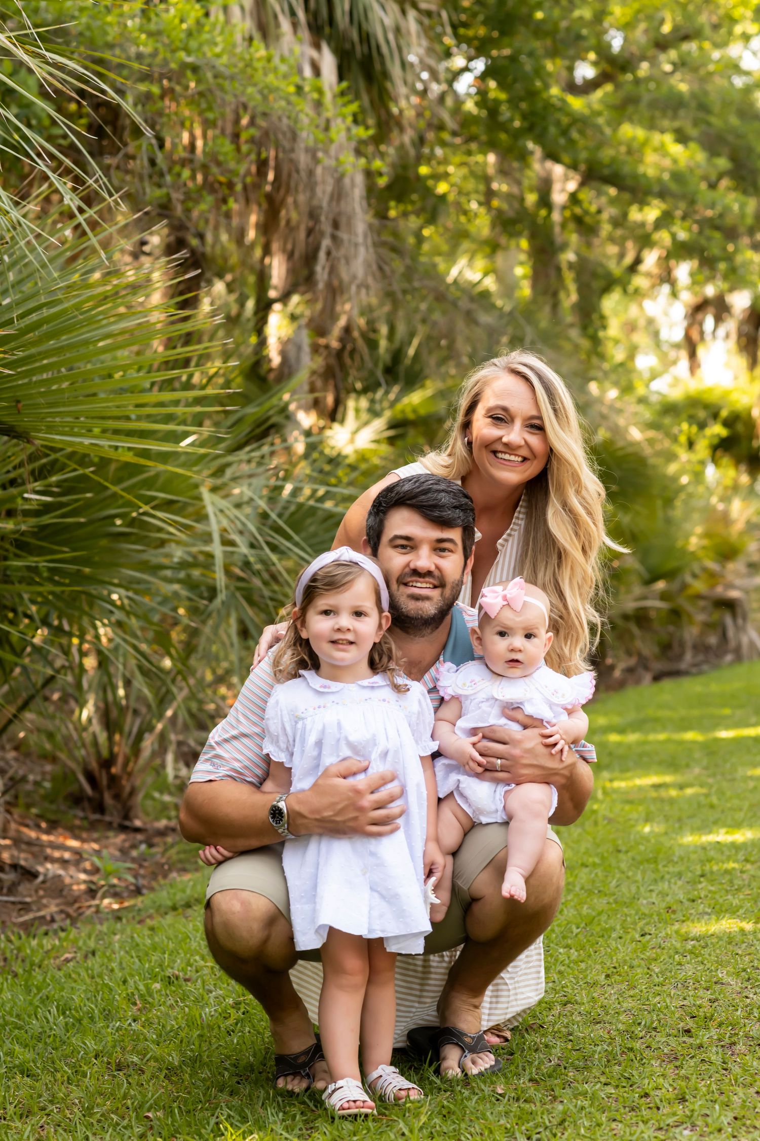 A family poses together on green grass with tropical palm trees in a sunny garden setting.