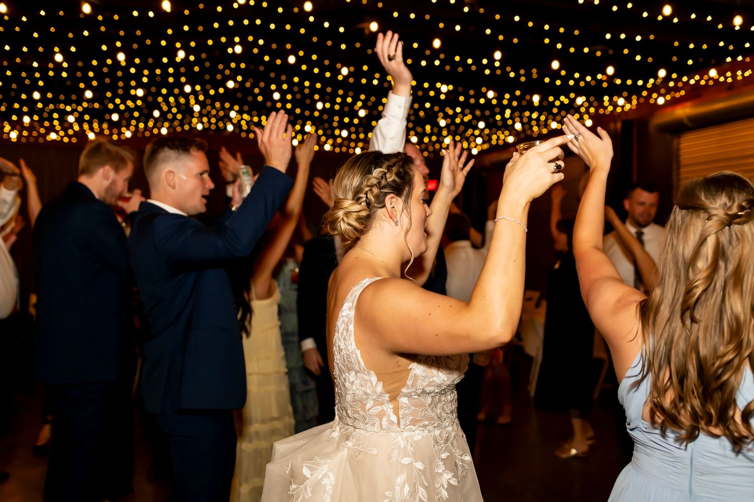 Wedding guests dance and celebrate under a ceiling of twinkling fairy lights at a reception.