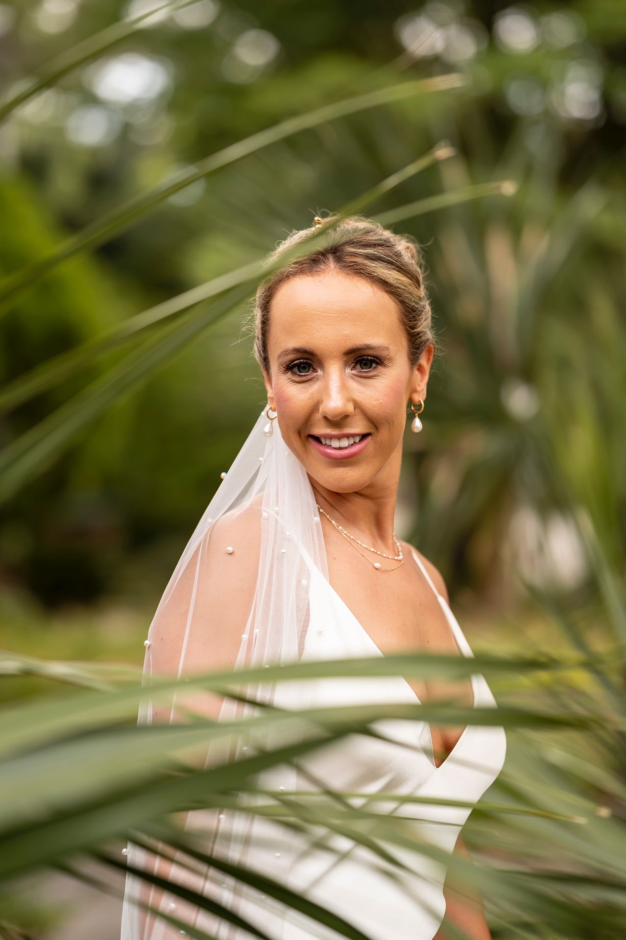 A bride in a white wedding dress and veil smiles through tropical palm leaves on her wedding day.