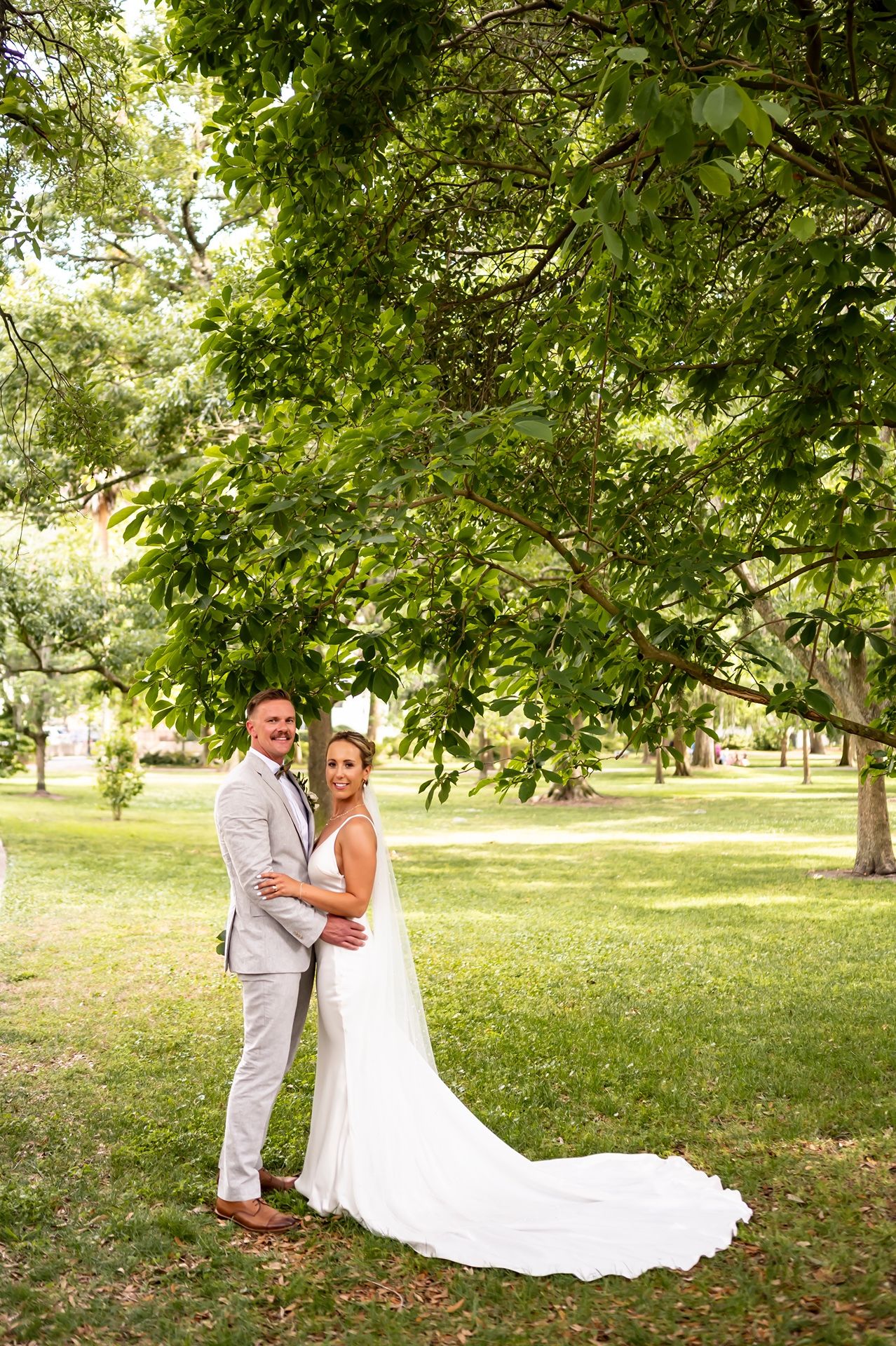 A couple in light-colored wedding attire embraces under lush green trees in a park setting.