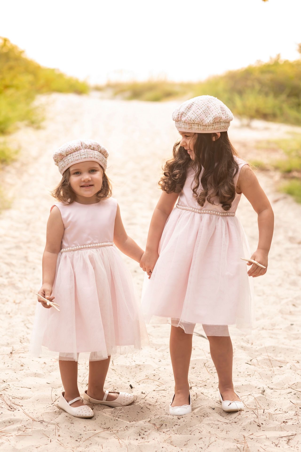 Two young children in matching pink dresses and berets hold hands while walking on a sandy path during golden hour.