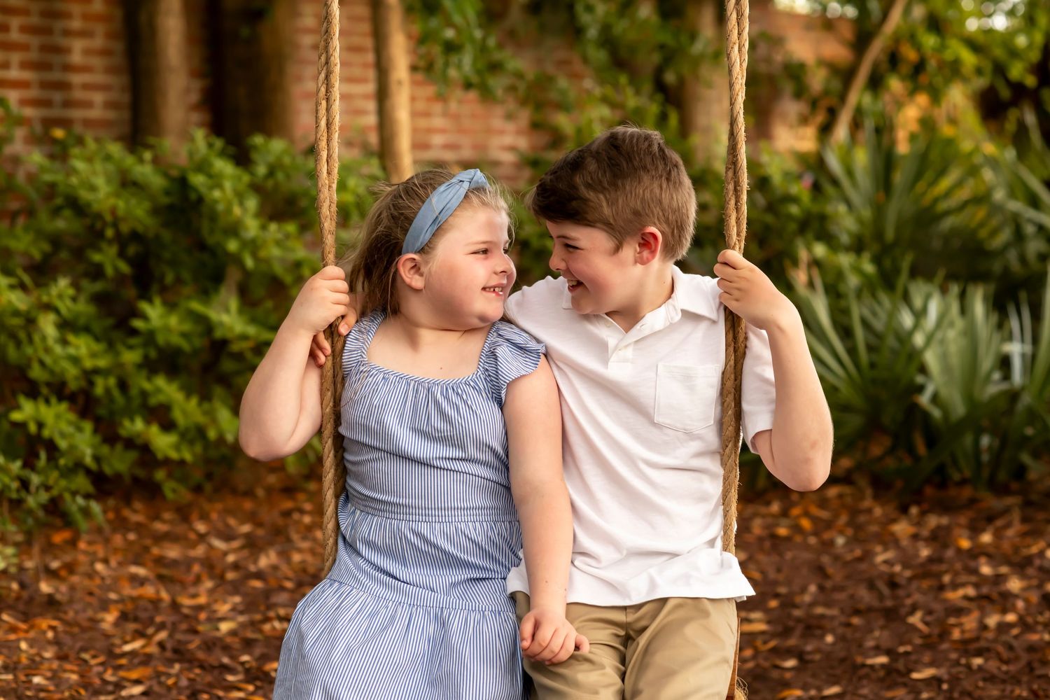 Two young children sit together on a swing in a leafy outdoor setting, smiling at each other.