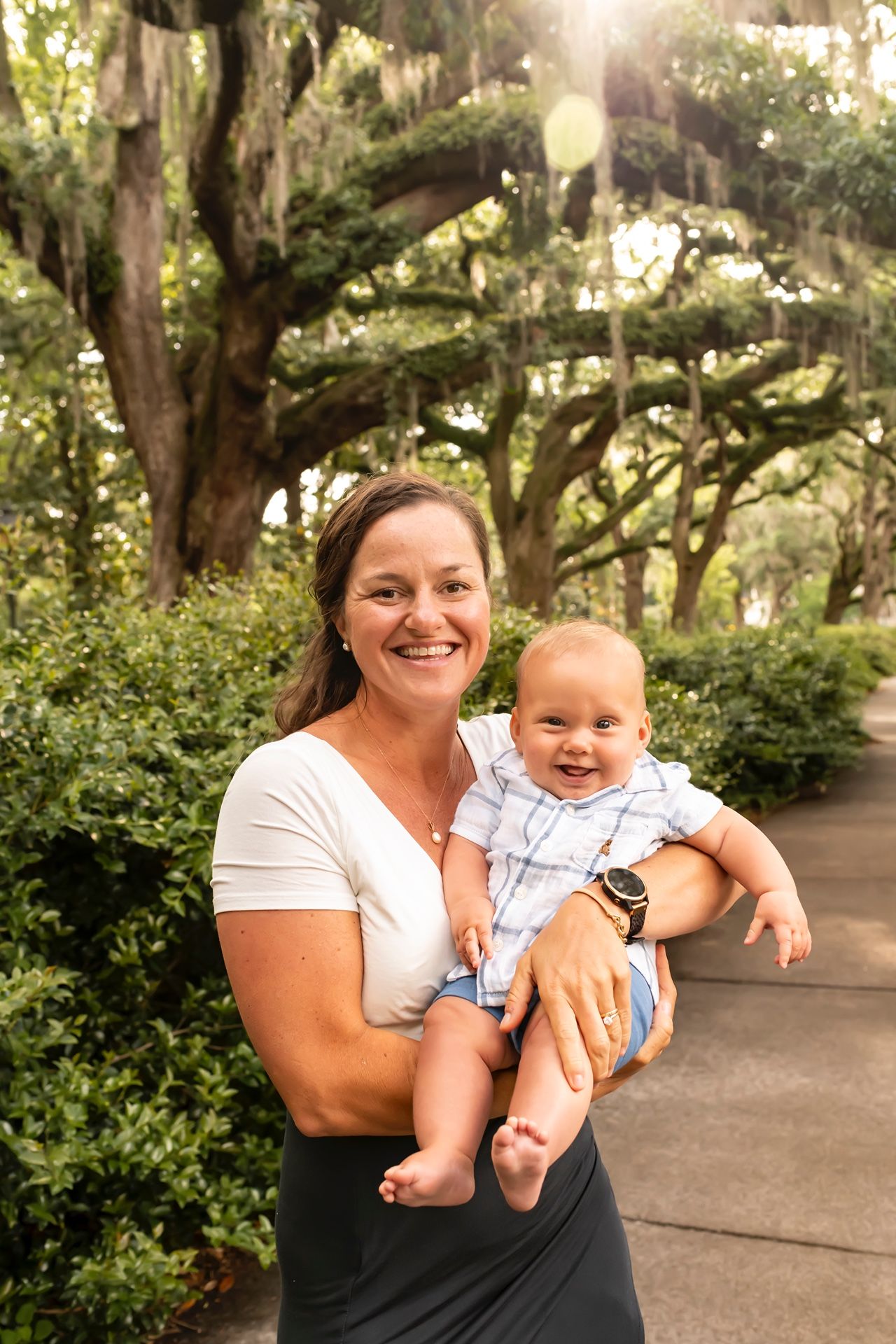 A joyful baby and caregiver share a sweet moment together under moss-draped oak trees in a park setting.